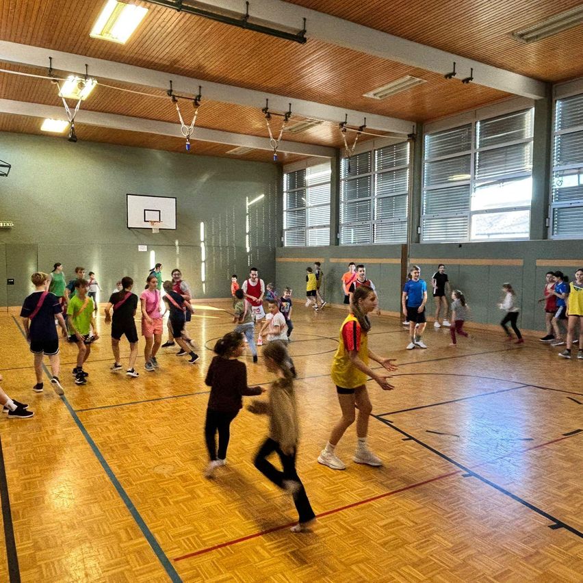 Eine Gruppe von Menschen spielt Basketball auf einem Hallenspielfeld. Einige Spieler tragen Turnschuhe, während andere barfuß sind. Sie laufen und lachen.