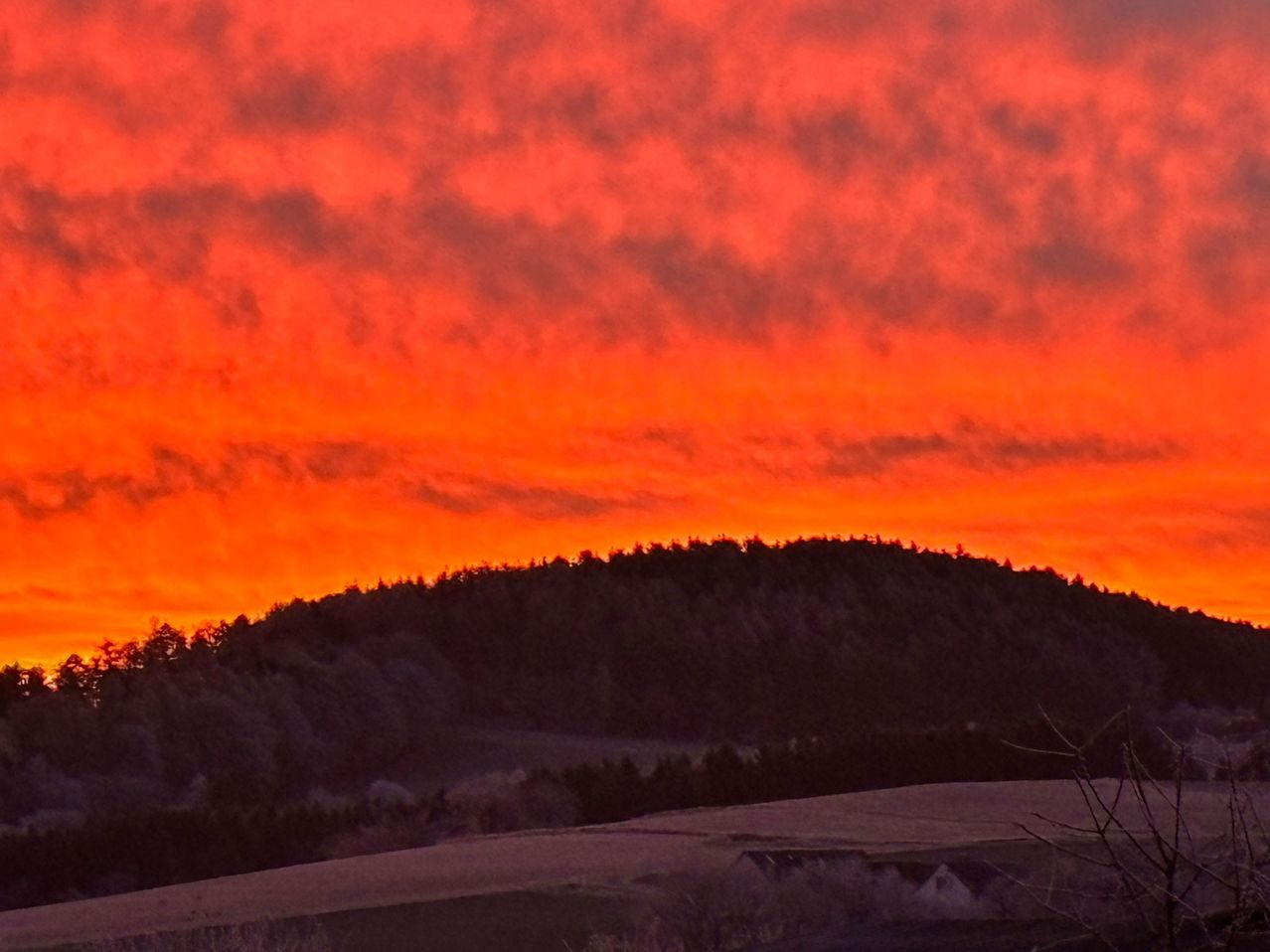 Ein lebhaftes Sonnenuntergehen mit orangen und rosa Farbtönen füllt den Himmel über einem silhouettierten Hügel und Wald.