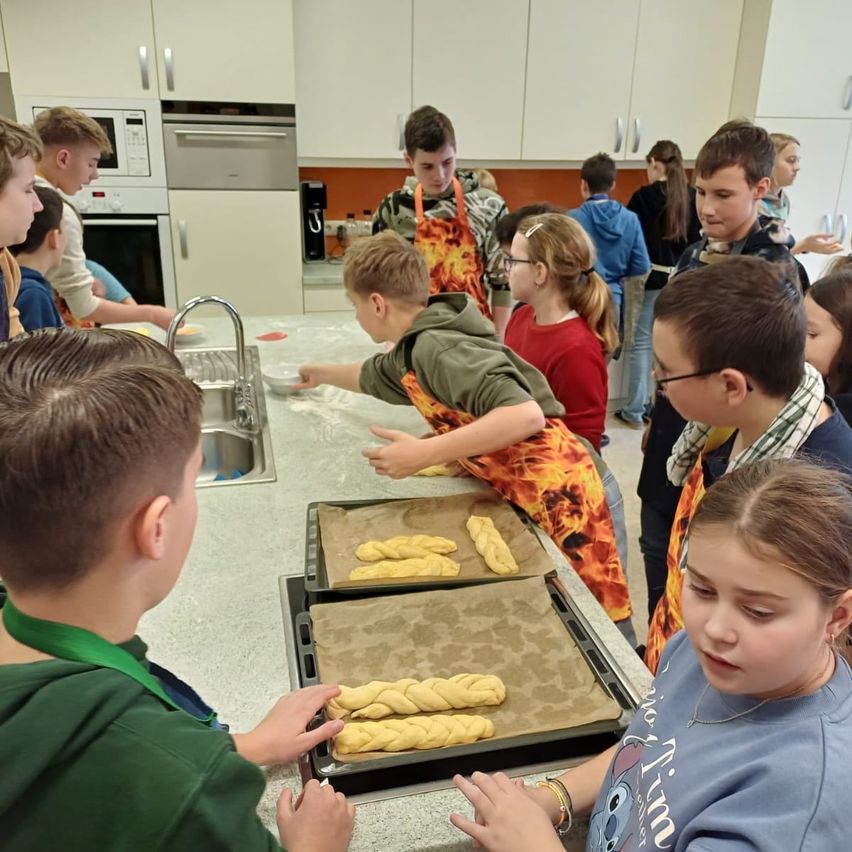 Eine Gruppe von Kindern ist in einer Küche und bereitet Essen zu. Einige kneten Teig, während andere zusehen. Tabletts mit gebratenem Teig sind auf dem Tisch.