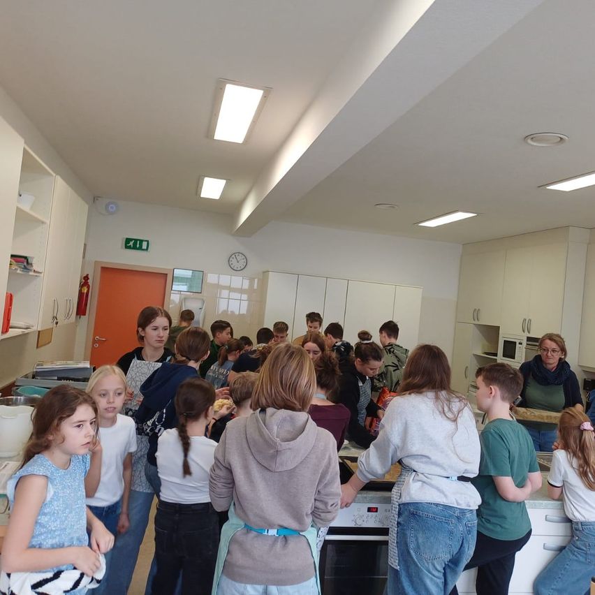 Eine Gruppe von Kindern und Erwachsenen versammelt sich in einer Küche, wahrscheinlich um Essen zuzubereiten. Einige tragen Schürzen, und eine Frau steht bei einer Mikrowelle. Andere versammeln sich um einen Tisch, und eine Uhr und ein Ausfahrtsschild befinden sich an der Wand.