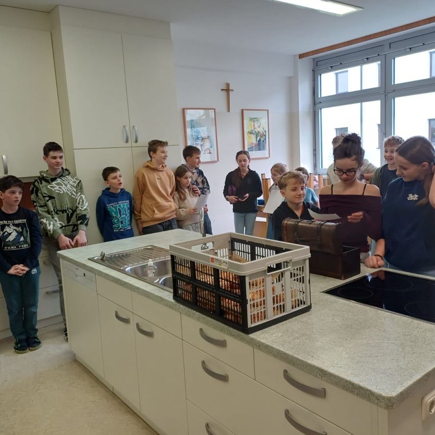 Mehrere Kinder und eine Frau stehen um einen Tresen herum und betrachten Eier in einem Korb. Der Tresen hat ein Waschbecken, einen Korb und eine Tasche. Ein Kreuz ist an der Wand und es gibt gerahmte Bilder und ein Fenster.
