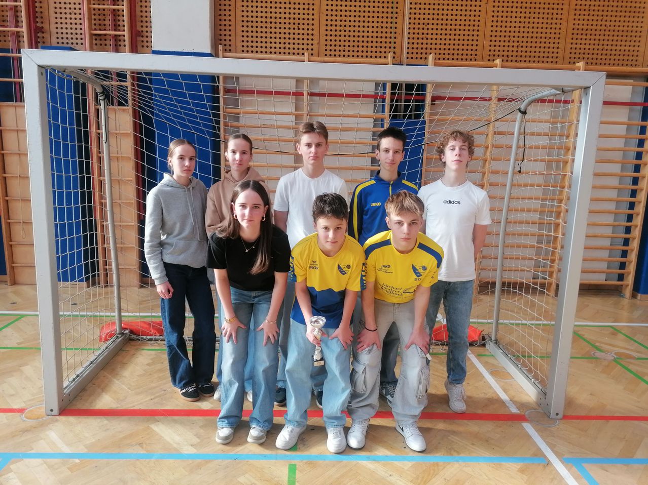 Eine Gruppe junger Menschen steht vor einem Sporttor und hält einen Pokal. Sie tragen verschiedenfarbige Shirts, und der Boden ist mit Linien markiert.