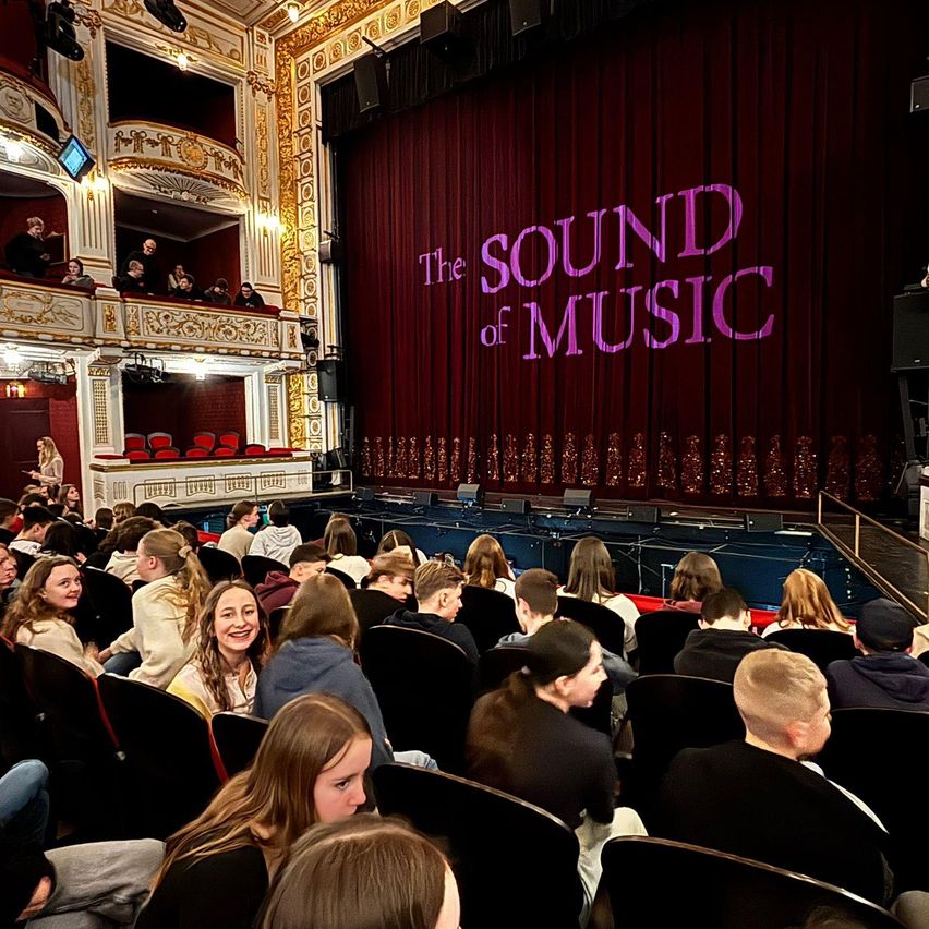 Publikum in Theaterstühlen, mit Blick auf die Bühne, auf der 'The Sound of Music' steht. Rote Samtvorhänge.