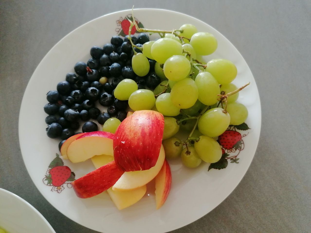 Ein weißer Teller mit Blumendesign zeigt verschiedene Früchte: grüne Trauben, Blaubeeren und Apfelscheiben.