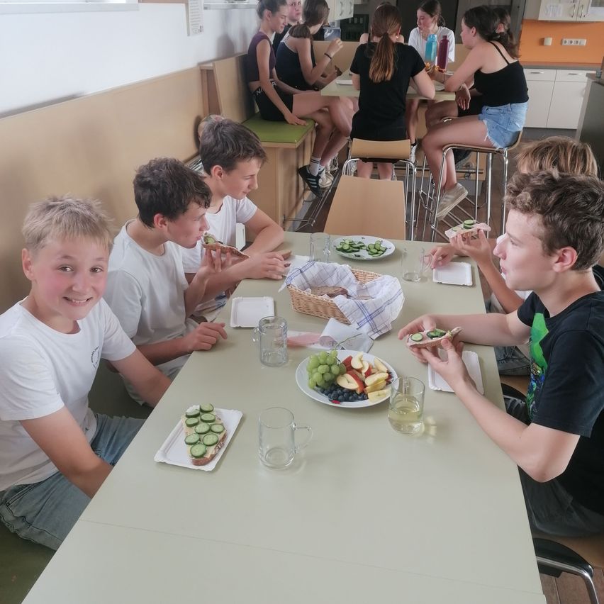 Eine Gruppe von Jungen und Mädchen isst an einem Tisch mit verschiedenen Speisen, einschließlich Obst und Sandwiches, in einem Essbereich.