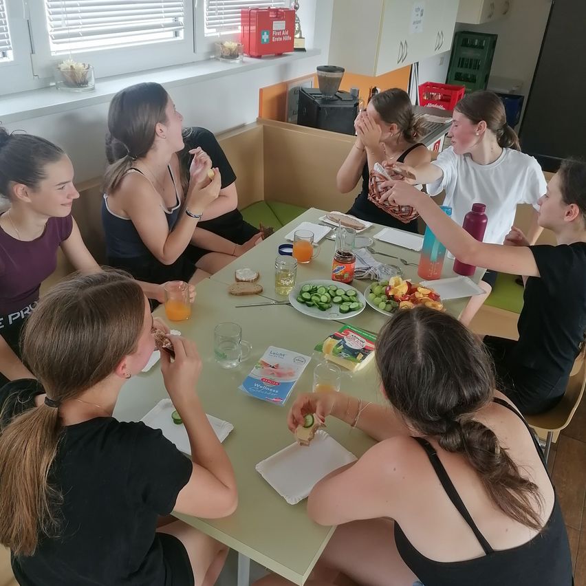 Eine Gruppe von Mädchen sitzt um einen Tisch und isst und trinkt. Einige Leute halten Essen. Auf dem Tisch sind eine Flasche, ein Becher und ein Teller.