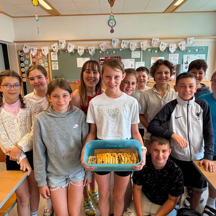 Gruppe von Kindern in einem Klassenzimmer lächelt mit einer Person, die eine Schale mit Bananenbrot hält. Sie sind von Dekorationen an der Wand umgeben.