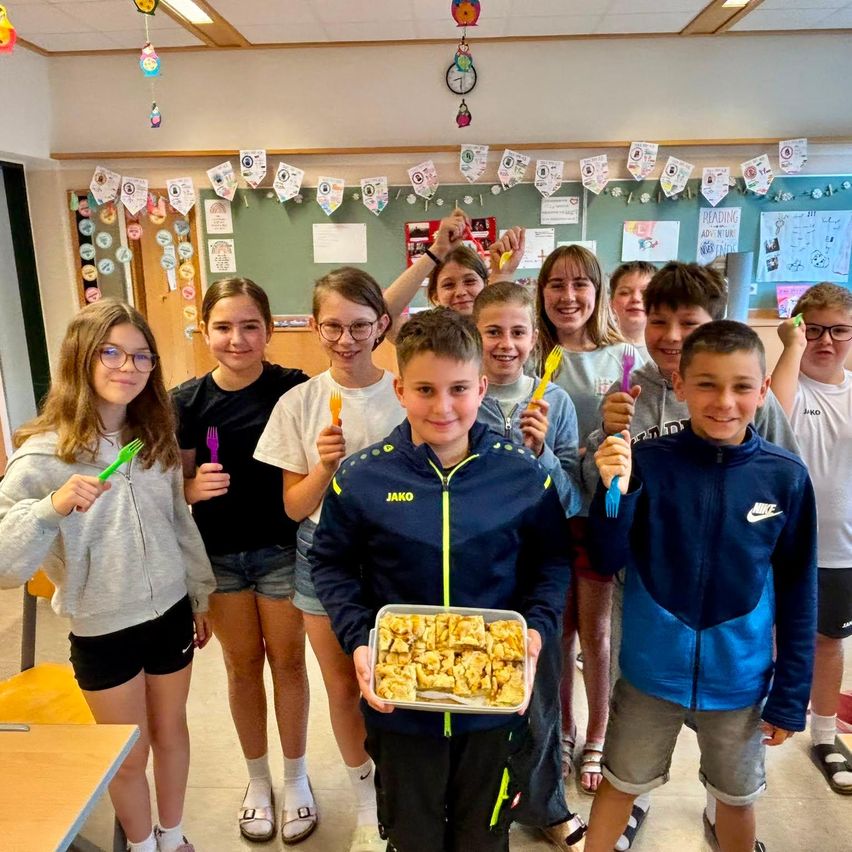 Eine Gruppe von Kindern in einem Klassenzimmer mit einem Lehrer, die Gabeln und eine Schale mit Essen halten. Das Klassenzimmer ist mit Papieren und einer Uhr an der Wand dekoriert.