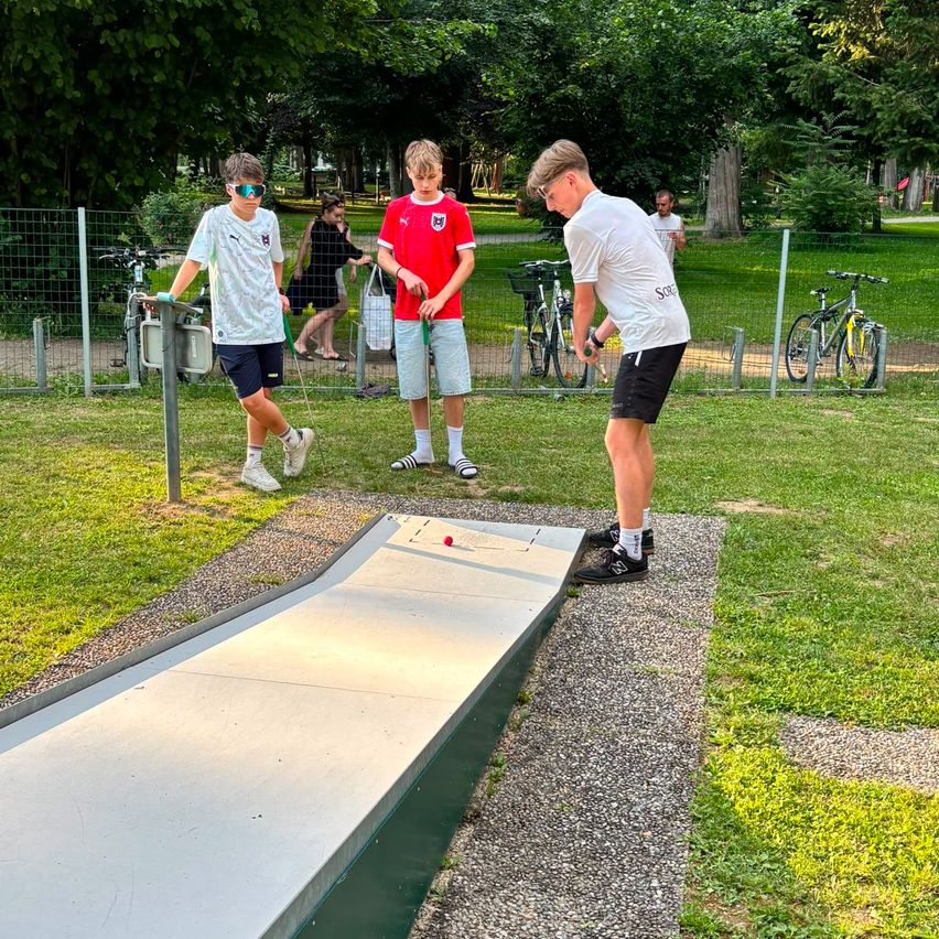 Drei Jungen spielen Minigolf an einem sonnigen Tag in einem Park. Ein Junge ist kurz davor, den Ball zu schlagen.