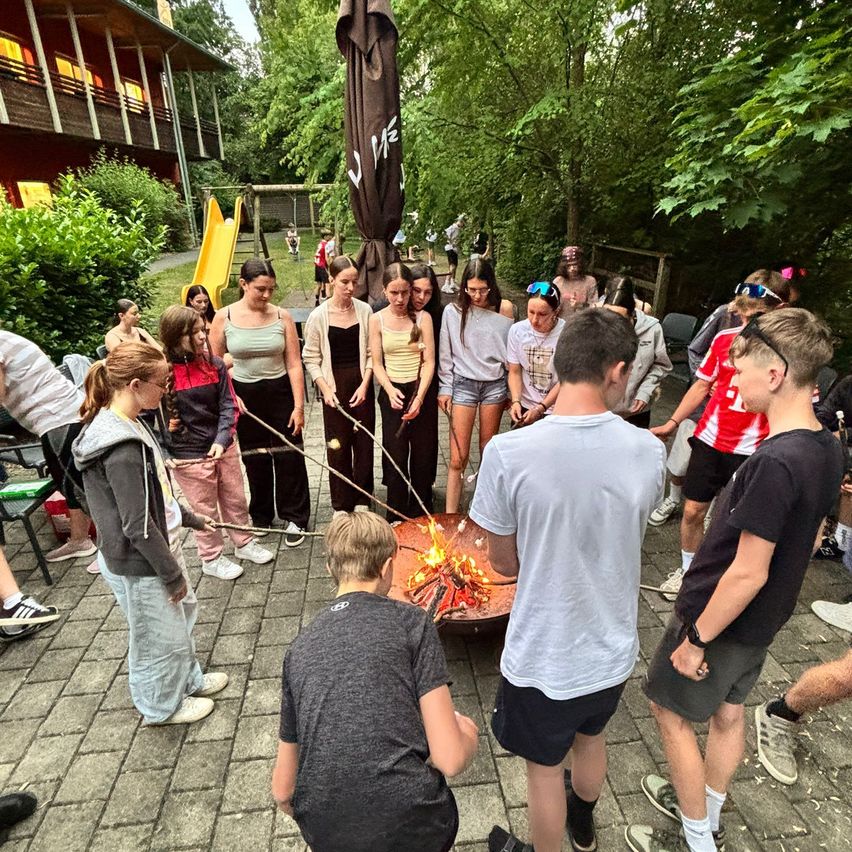 Eine Gruppe von Teenagern ist um ein Lagerfeuer in einem Außenbereich versammelt. Sie halten Stöcke mit Marshmallows zum Rösten. Einige stehen, andere sitzen auf dem Boden. Die Szene ist von Bäumen und einem Gebäude umgeben.