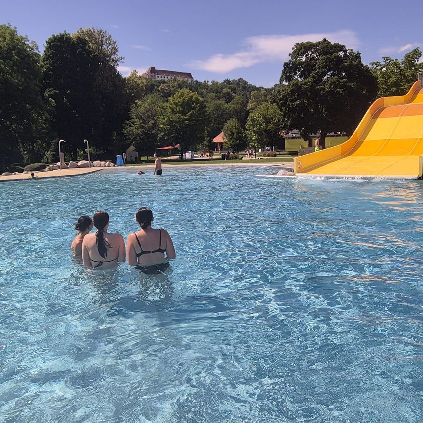 Drei Frauen stehen in einem Pool mit einem gelben Wasserrutsche im Hintergrund. Bäume und ein Gebäude sind in der Ferne sichtbar.