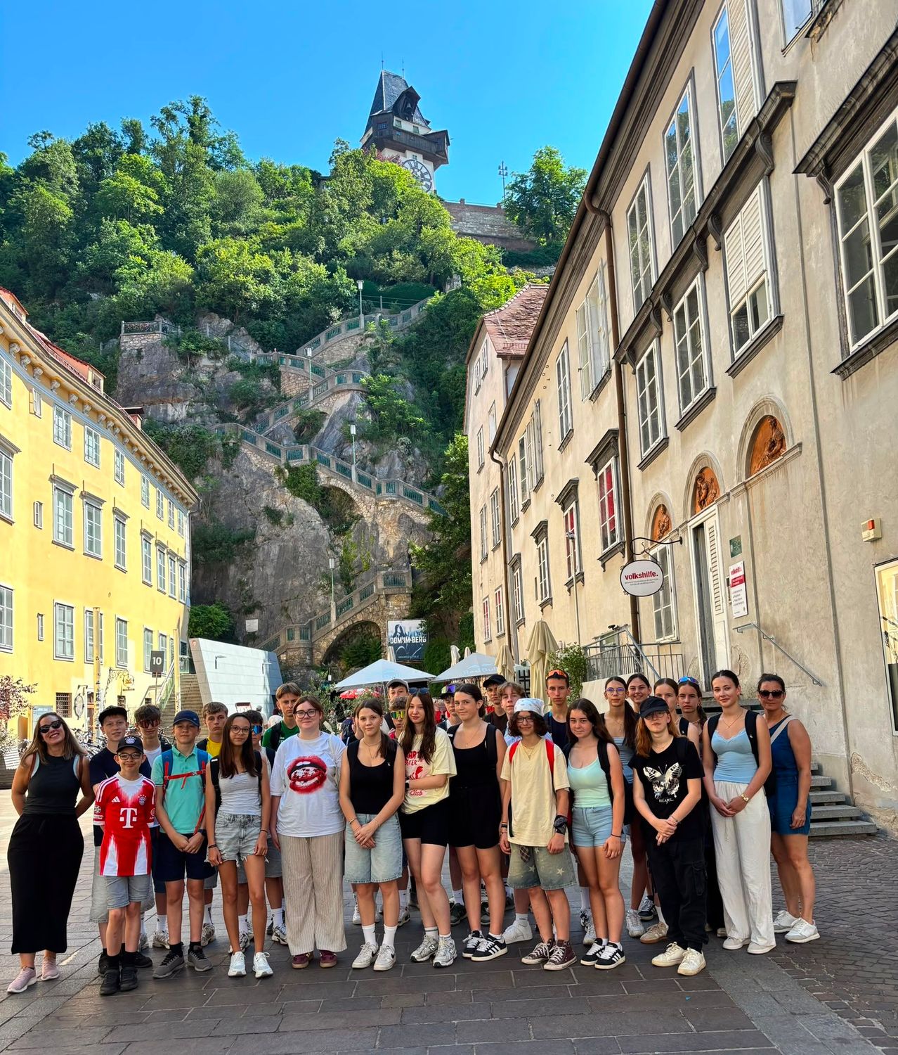 Eine Gruppe von Menschen posiert für ein Foto vor Gebäuden und einer Burg auf einem Hügel mit Treppen und Grünflächen.