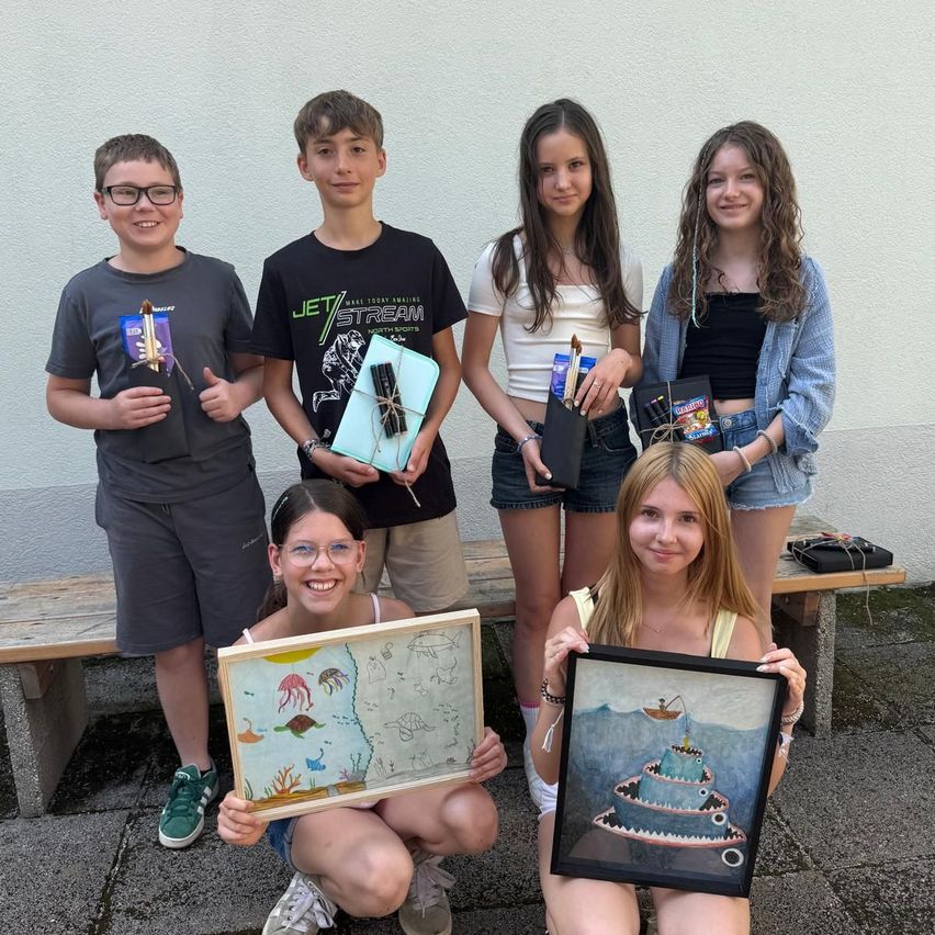 Sechs junge Künstler posieren mit ihren Kunstwerken vor einer Wand. Zwei halten gerahmte Gemälde und die anderen halten Zeichnungen. Sie alle lächeln und zeigen ein Gefühl der Errungenschaft.