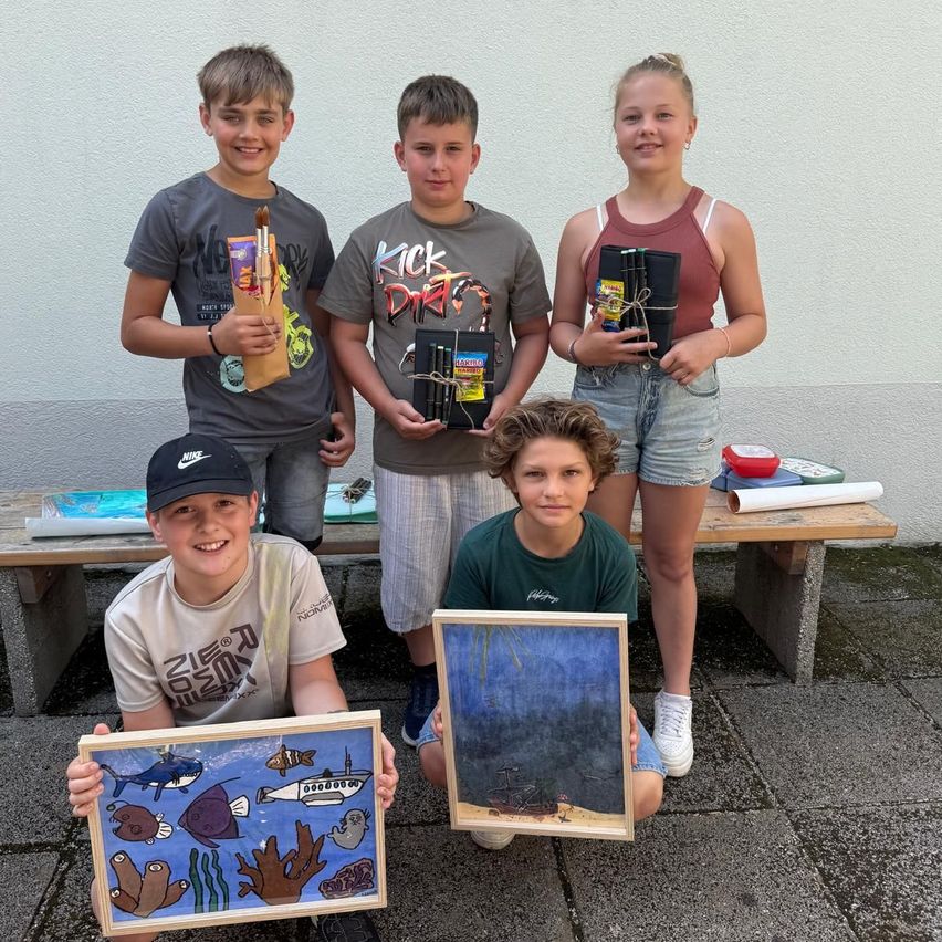 Fünf Kinder stehen draußen zusammen und halten Malutensilien. Sie posieren mit ihren Zeichnungen. Eines hat ein U-Boot, ein anderes eine Meeresszene. Sie lächeln für die Kamera.