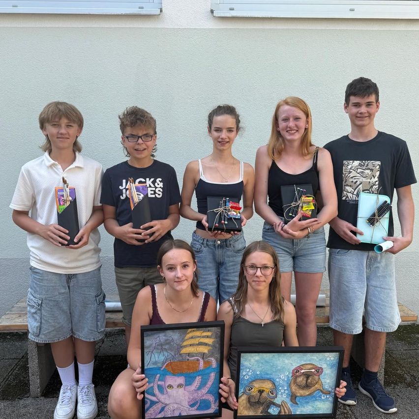 Sieben junge Leute halten Kunstzubehör und Gemälde in den Händen. Die Gruppe steht vor einem Gebäude, zwei Mädchen knien und zwei Jungen sitzen auf einer Bank.