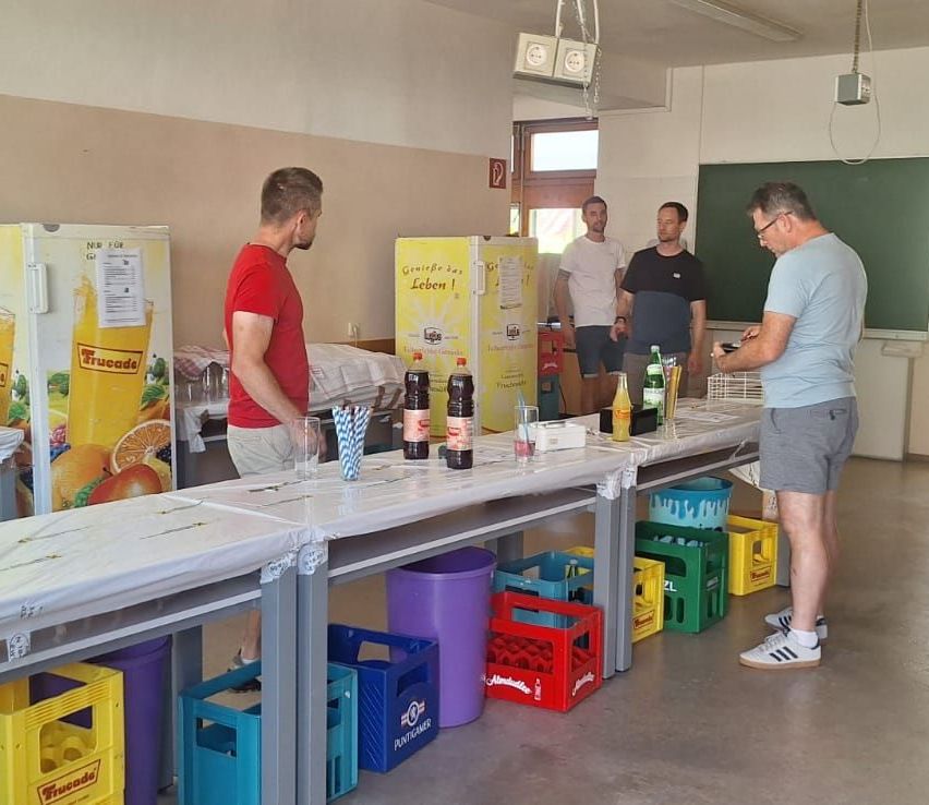 Eine Gruppe von Männern ist in einem Raum mit einem langen Tisch und mehreren Gegenständen darauf. Ein Mann schaut auf sein Handy, während ein anderer Mann ihn ansieht.