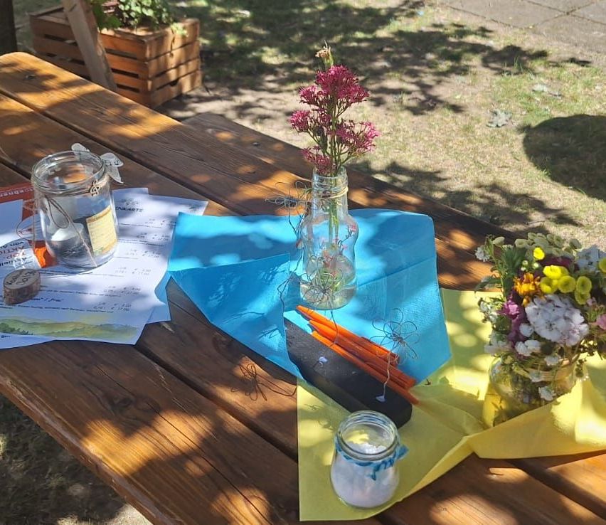 Ein Holz-Picknicktisch hat ein blaues Tuch mit Blumen und einem Glas. Ein gelbes Tuch darunter mit einem Stein, Stöcken und einem Glas. Papiere sind links.