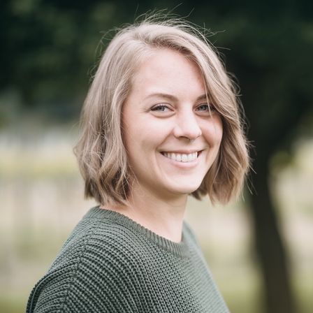 Eine Frau mit blonden Haaren lächelt in einer Außenumgebung. Sie trägt einen grünen gestrickten Pullover und steht vor einem unscharfen Baum.