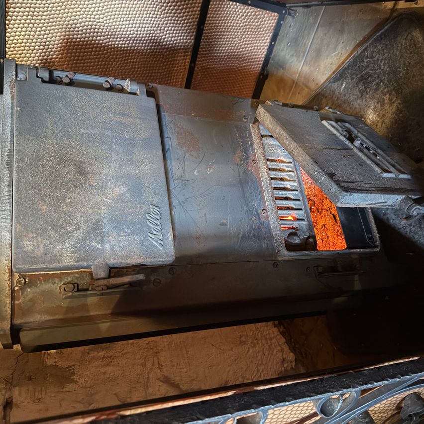 Das Bild zeigt einen alten, rostigen Ofen mit dem Wort Navien darauf. Der Ofen hat eine offene Tür, durch die ein brennendes oranges Feuer im Inneren zu sehen ist. Er steht auf einem Steinboden.