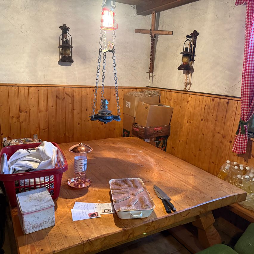 Ein hölzerner Esstisch hat verschiedene Gegenstände darauf, darunter ein Korb mit Kleidung, eine Lampe, ein Tablett und ein Messer. Hinter dem Tisch befinden sich Holzwände mit hängenden Laternen, Kisten und einem Kreuz.