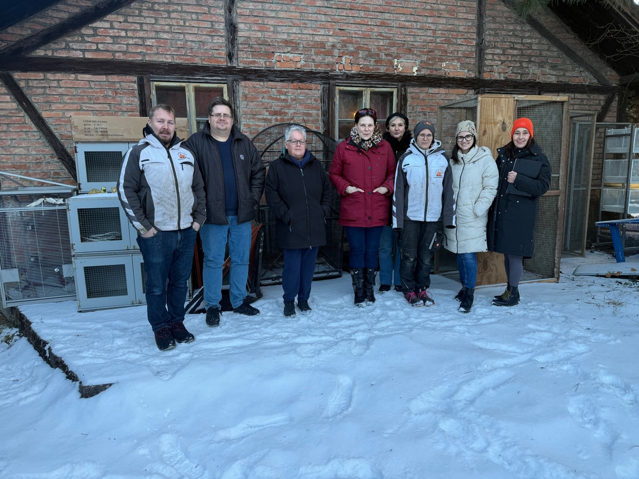 Eine Gruppe von Menschen steht im Schnee vor einem Backsteingebäude. Sie sind alle warm gekleidet. Es gibt zwei Frauen, die eine Brille und Mützen tragen, und eine Frau in der Mitte mit einem Schal. Hinter ihnen befinden sich einige Käfige.
