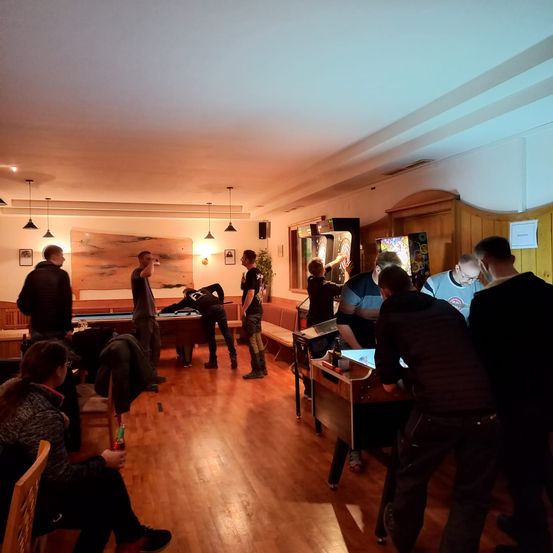 Eine Gruppe von Menschen befindet sich in einem Raum mit Holzböden und Arcade-Spielen. Einige spielen Tischspiele, andere schauen zu.