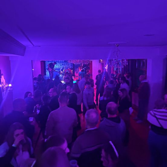 Eine Band spielt auf der Bühne unter lila Licht in einem schwach beleuchteten Raum, vor einer Menge tanzender und zuschauender Menschen.