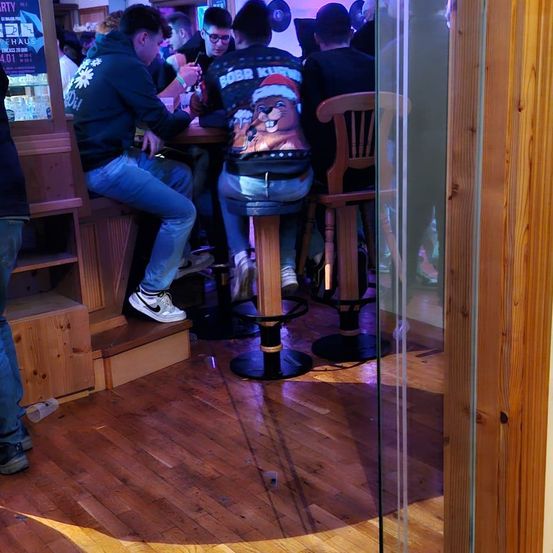 Eine Gruppe von Menschen sitzt an einer Bar, einer trägt ein Weihnachtspullover. Ein anderer trägt eine Brille und Turnschuhe. Sie sind von Holzmöbeln umgeben.