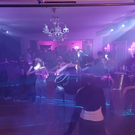 Eine Gruppe von Menschen tanzt in einem Nachtclub mit lila Lichtern und einer Kristallleuchte. Eine Frau in einem schwarzen Shirt hält ein Handy.