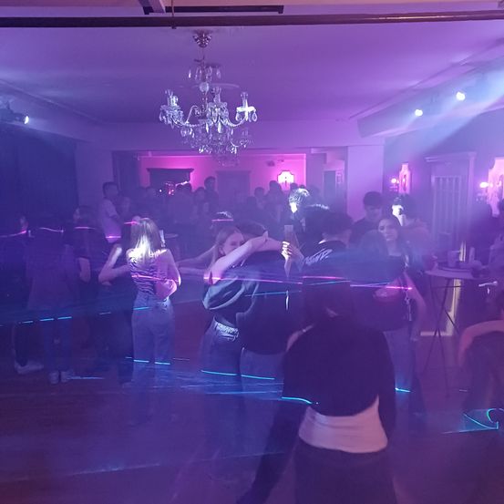 Eine Gruppe von Menschen tanzt in einem Nachtclub mit lila Lichtern und einer Kristallleuchte. Eine Frau in einem schwarzen Shirt hält ein Handy.