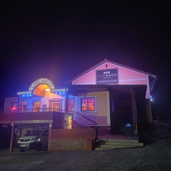 Live Haus, ein lebhaftes Musiklokal, steht nachts beleuchtet. Das Gebäude hat rosa Wände und einen Balkon mit Geländern. Ein Auto ist davor geparkt.