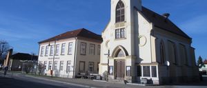 Eine weiße Kirche mit Turm und Uhr, neben einem beigen Schulgebäude, unter einem klaren blauen Himmel.