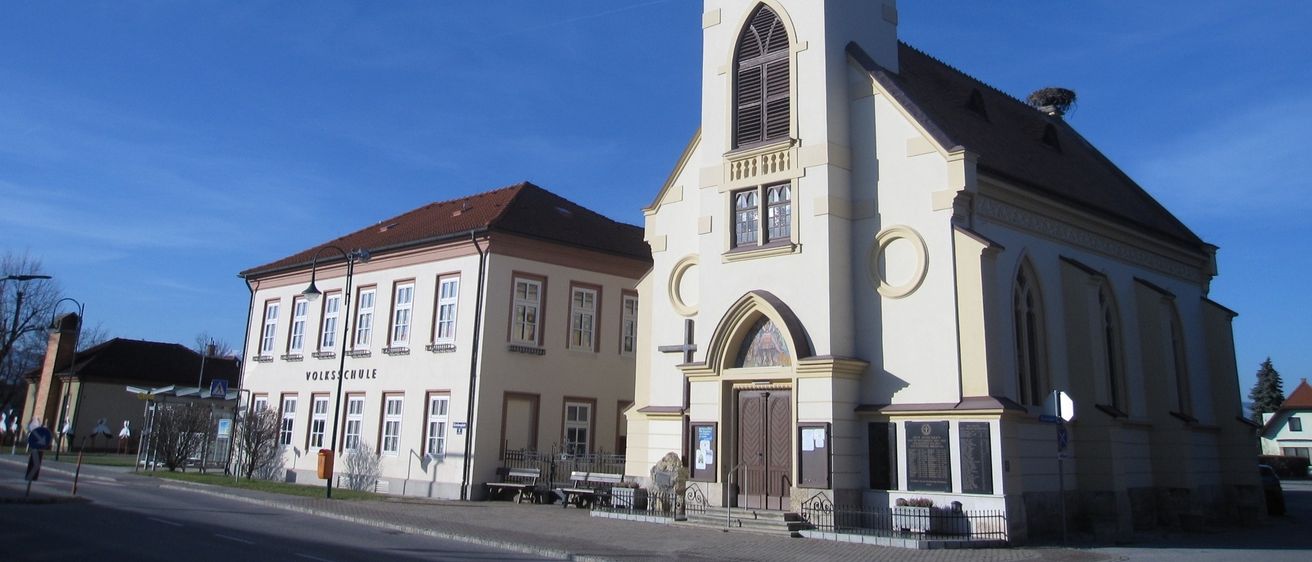 Eine weiße Kirche mit Turm und Uhr, neben einem beigen Schulgebäude, unter einem klaren blauen Himmel.