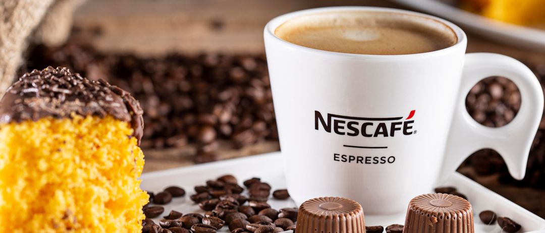 Eine Nescafe Espresso-Tasse mit einem schokoladenüberzogenen Kuchen und Schokoladentrüffeln auf einem weißen Teller, umgeben von Kaffeebohnen.