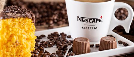 A white plate has a Nescafe Espresso cup, a yellow cake, chocolate truffles, and coffee beans on a wooden surface.