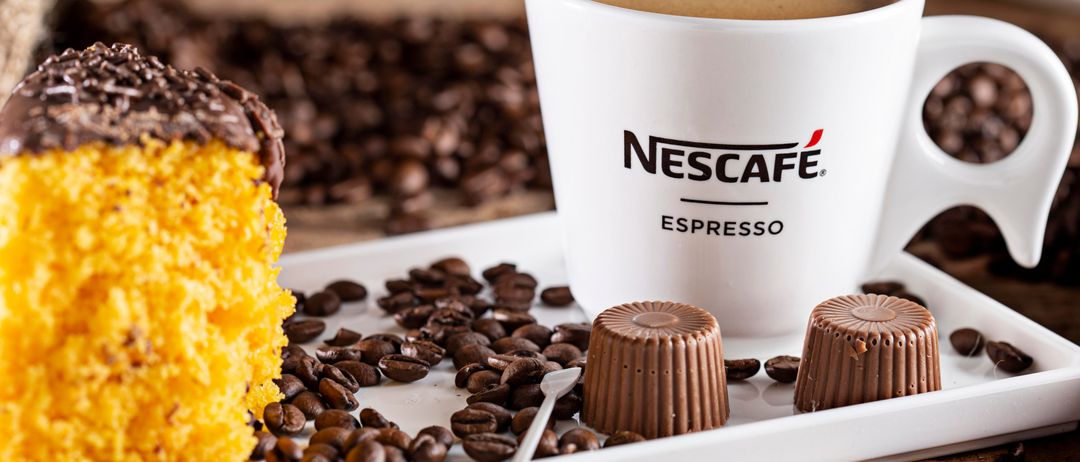 Ein weißer Teller hat eine Nescafe Espresso-Tasse, einen gelben Kuchen, Schokoladentrüffel und Kaffeebohnen auf einer Holzoberfläche.