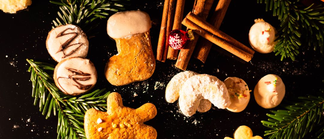 Eine Auswahl an Weihnachtsplätzchen, einschließlich Lebkuchenmännern und einem stiefelförmigen Keks. Die Kekse sind von Zimtstangen, Tannenzweigen und einer roten Weihnachtskugel umgeben.