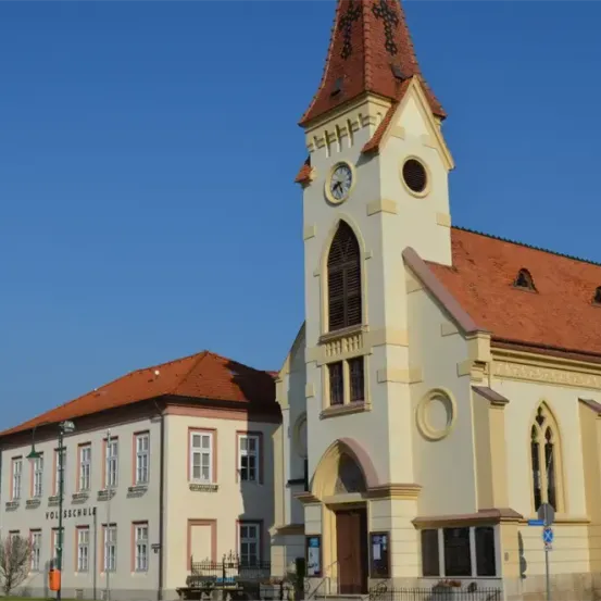 Ein architektonisches Bild einer Kirche mit Glockenturm und rotem Dach. Die Kirche ist weiß und hat bogenförmige Fenster. Ein Gebäude mit rotem Dach befindet sich neben ihr.