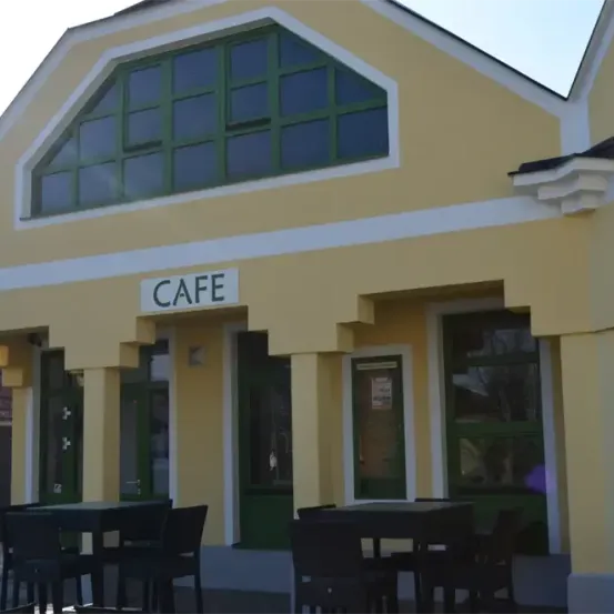 Das gelbe Cafe-Gebäude hat große Fenster und eine Glastür. Es gibt Tische und Stühle draußen. Das Wort CAFE steht oben.