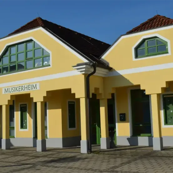 Das Äußere eines leuchtend gelben Gebäudes, mit der Aufschrift 'Musikheim', verfügt über mehrere Fenster, ein schwarzes Dach und grüne Akzente.