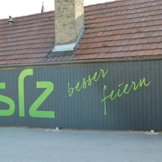 Ein Gebäude mit einem roten Dach und einem Schornstein, mit einem grünen Schild, das den Text 'sfz besser feuern' auf einer grauen Wand zeigt.