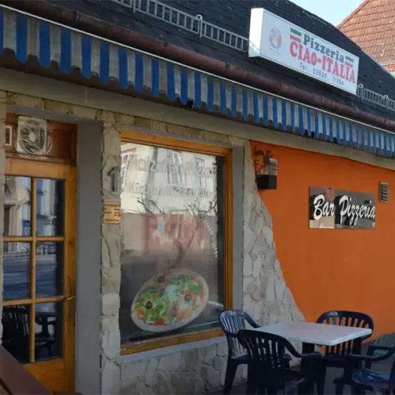 Eine Pizzeria namens Ciao-Italia mit einem Tisch und Stühlen draußen. Das Fenster zeigt eine Pizza auf einem Teller. Das Schild darüber hat weißen Text auf blau-weißem Hintergrund.