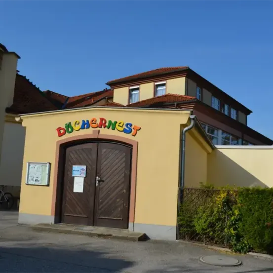 Ein hellgelbes Gebäude mit dem Wort Dochernest in Regenbogenfarben über dem Eingang geschrieben. Eine braune Holztür ist offen. Hinter dem Gebäude befindet sich ein Haus mit einem roten Dach. Ein Fahrrad ist auf der linken Seite geparkt.