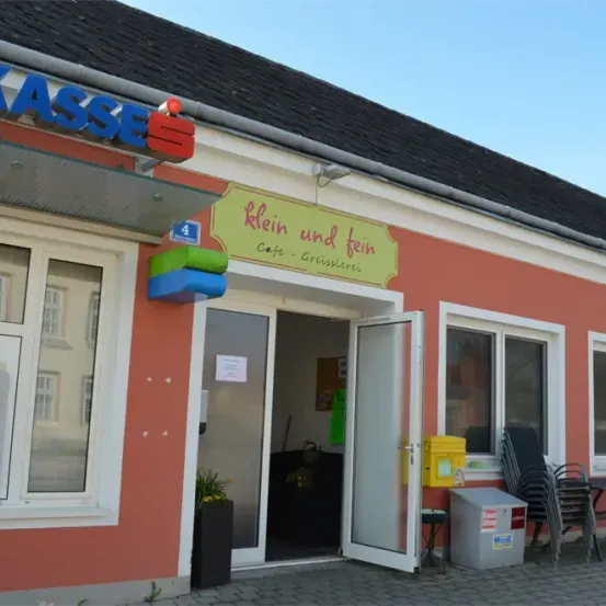 Der Eingang eines Cafes mit einem gelben Schild, auf dem Klein und Fein Cafe steht. Das Cafe hat Glastüren und Fenster. Ein Topf mit Pflanzen steht vor dem Eingang. Stühle und ein Tisch sind an der Seite.