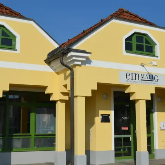 Ein gelbes Gebäude mit grünen Fenstern und einem Schild, das 'einMALIG Friseurmeister & Makeup Artist' besagt.