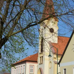 Eine alte Kirche mit einem roten Dach und einem hohen Turm steht in einer Stadt mit klarem Himmel darüber. Sie hat bogenförmige Fenster und einen Glockenturm. Eine Straßenlaterne steht an der Seite.
