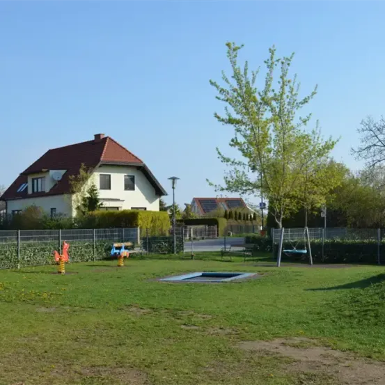 Ein Haus mit einem roten Dach und einem Zaun davor. Ein Spielplatz mit Schaukel, Rutsche und einem Pool. Bäume und Büsche sind in der Umgebung.