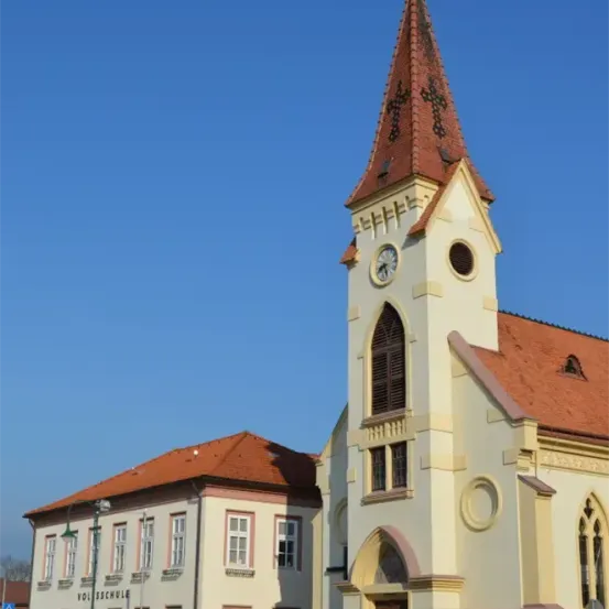 Eine Kirche mit einem hohen Turm und einer Uhr, unter einem klaren blauen Himmel. Das Gebäude hat ein rotes Dach und gelbe Wände. In der Nähe befindet sich ein weißes Gebäude mit mehreren Fenstern, auf dem 'Vollschule' steht.