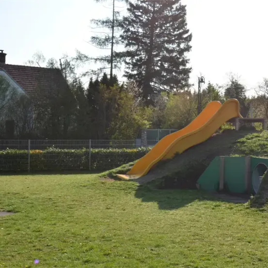 Ein sonniger Tag auf einem Spielplatz mit einer gelben Rutsche und einem grünen Tunnel im Gras in der Nähe eines Hauses und Bäumen.
