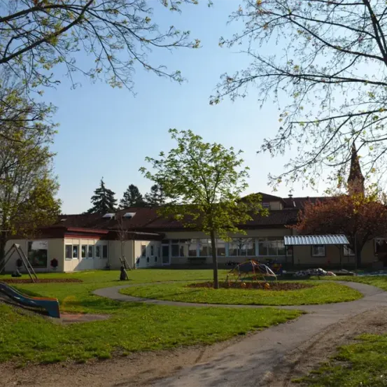 Ein Schulgebäude mit braunem Dach und mehreren Fenstern ist von Rasen und Bäumen umgeben. Ein Rutschvergnügen und Spielgeräte sind im Hof.